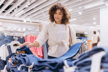 Smiling young woman shopping for jeans in a clothing store, holding a pair of denim trousers and looking at other clothes on hangers