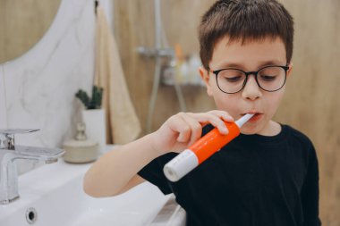 Concentrated little boy brushing teeth with modern electric toothbrush while standing in light bathroom at home