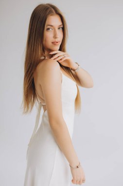 Young woman with long brown hair wearing elegant white dress and jewelry posing in studio on white background