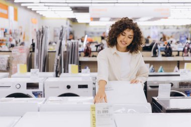 Customer comparing prices and features of washing machines while shopping in an electronics store, making informed decisions for home needs
