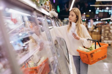 Müşteri marketten sebze seçiyor alışveriş listesi okuyor, elinde taze sebzelerle turuncu sepet tutuyor.