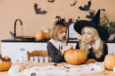 Mother wearing a witch hat and daughter joyfully carving pumpkins together for a festive Halloween party at home, creating lasting memories