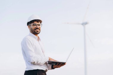 Young Arabian engineer with hardhat and laptop working near a wind turbine for sustainable energy production