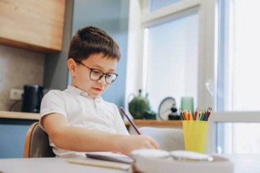 Concentrated young boy wearing eyeglasses drawing with colored pencils while sitting at table in cozy kitchen at home