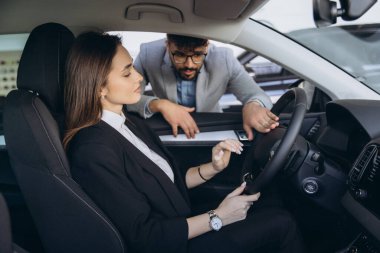 Salesman highlighting features of a new car while businesswoman sits in the driver's seat, engaged in conversation at the dealership