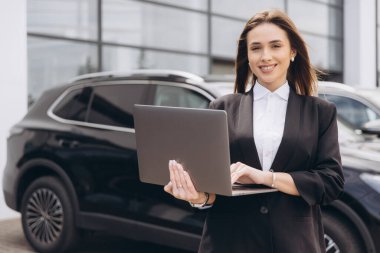 Saleswoman smiling and using laptop in front of a car in a car dealership