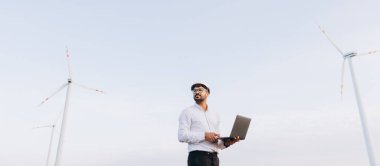 Arabian engineer with laptop and white helmet working in a wind turbine power plant for sustainable energy