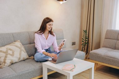 Smiling woman sitting comfortably on sofa using laptop and smartphone, enjoying benefits of working remotely in cozy home environment