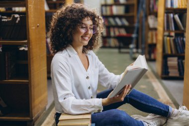 Young curly haired student wearing eyeglasses reading a book sitting on the floor in the library between bookshelves