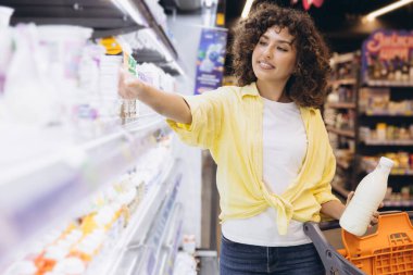 Smiling woman selecting yogurt from refrigerated shelves while holding milk bottle and shopping basket in supermarket