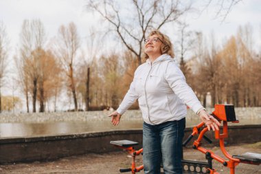Elderly woman enjoying exercising with outdoor gym equipment in a park, promoting healthy lifestyle and active aging