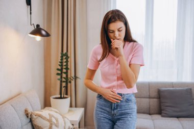 Sick woman coughing and experiencing abdominal pain while standing in her living room, surrounded by furniture and plants