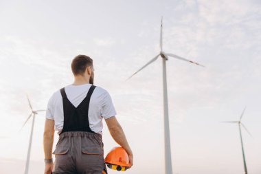 Wind turbine technician holding an orange helmet while gazing at multiple wind turbines in a vast renewable energy landscape