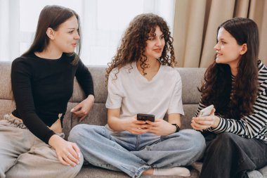 Three teenage girls are using their smartphones and talking, sitting on a comfortable sofa in the living room at home