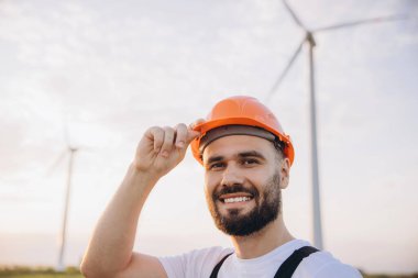 Smiling engineer adjusting his helmet while standing in front of wind turbines, showcasing a commitment to renewable energy and sustainability