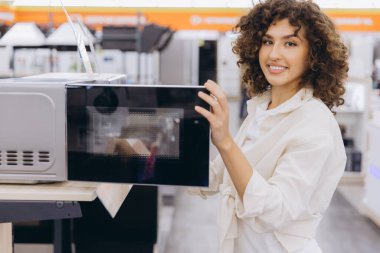 Smiling curly haired woman opening microwave oven door in home appliance store, choosing kitchen equipment