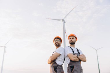 Two engineers with crossed arms standing back to back in a wind turbine farm, looking away, wearing hard hats and work overalls