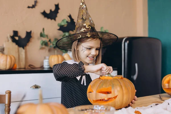Smiling little girl wearing a witch hat, joyfully carving a pumpkin in preparation for a fun Halloween party celebration at home