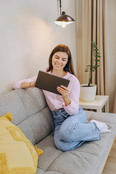 Young woman relaxing on comfortable sofa using digital tablet, enjoying online entertainment or working remotely from home