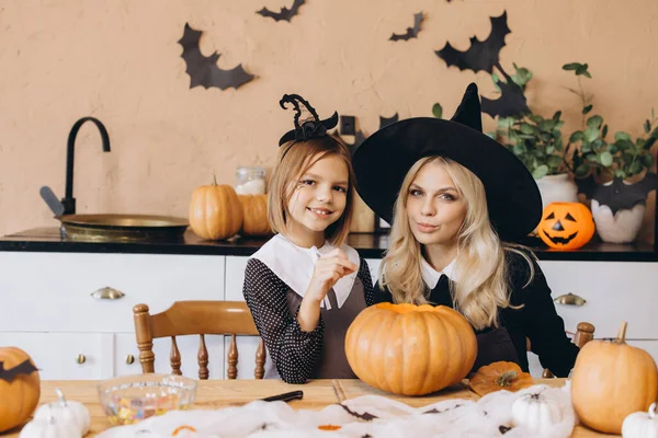 Mother and daughter wearing witch costumes, carving a pumpkin to make a jack o' lantern in a kitchen decorated for Halloween