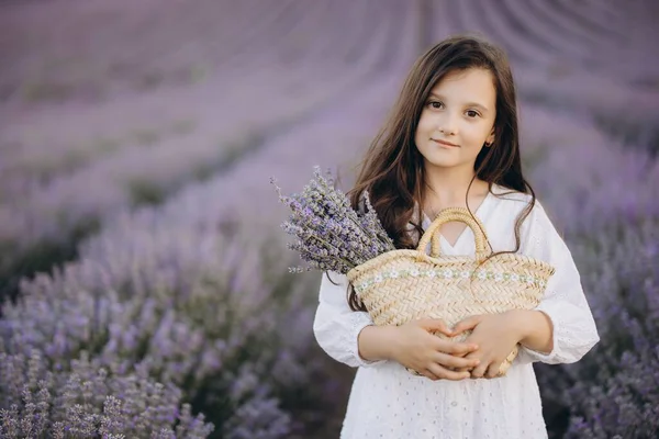 Girl wearing a flowing white dress, holding a wicker bag and a vibrant lavender bouquet while wandering through a picturesque lavender field