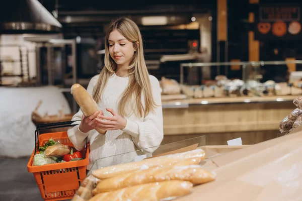 Müşteri bir bakkal dükkanından taze ekmek seçiyor, elinde renkli sebzelerle dolu bir alışveriş sepeti tutuyor ve