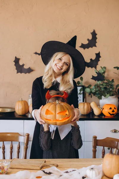 Mother wearing witch costume and daughter with devil horns holding carved pumpkin with candle inside during Halloween preparation