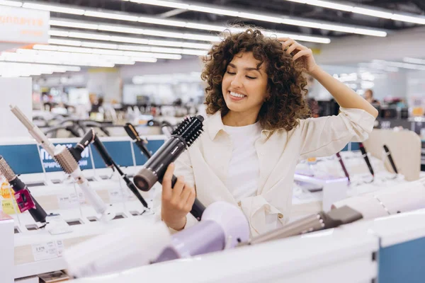 Smiling curly haired woman touching her hair and choosing a new hairdryer in an electronics store