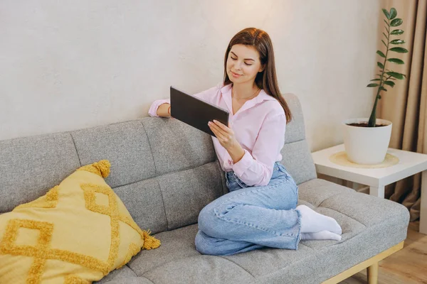 Woman sitting comfortably on a sofa, browsing the internet on a digital tablet in a cozy and inviting living room filled with natural light