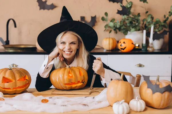Young woman wearing witch costume carving pumpkins for Halloween party, holding knife and looking mischievous
