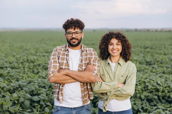 Interracial team of two agronomists standing confidently with arms crossed, posing in a lush, cultivated soybean field under a clear summer sky
