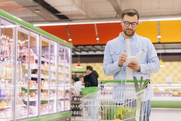 Focused customer taking notes in his notepad while checking shopping list near refrigerator with frozen food in supermarket