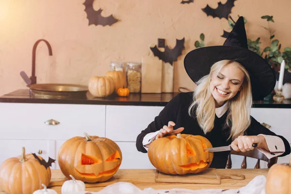 Young woman wearing a witch hat, happily carving pumpkins for a Halloween party in a festively decorated kitchen filled with autumn vibes