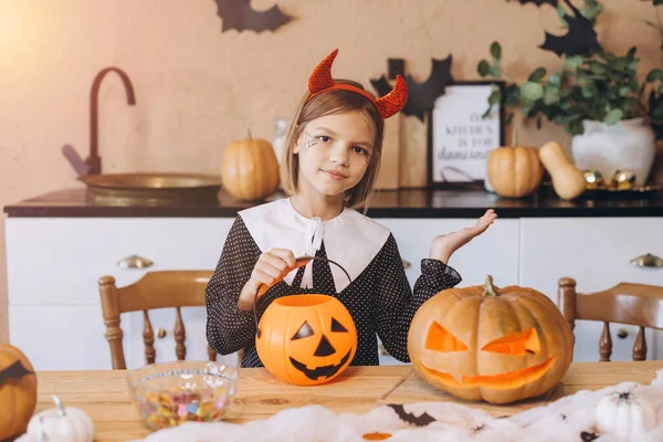 Girl wearing devil horns headband and halloween costume preparing a pumpkin basket with candy, sitting at a wooden table with carved pumpkins