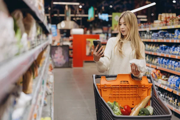 Young woman browsing her smartphone while shopping for groceries in a supermarket, selecting fresh produce and comparing prices