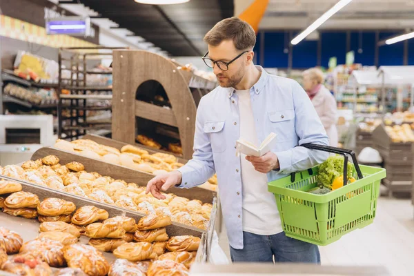 Man choosing pastry from a shelf, holding a shopping basket and a grocery list in supermarket bakery