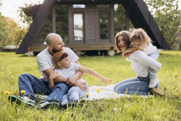Smiling family having fun sitting on blanket in the grass in front of their modern a frame house