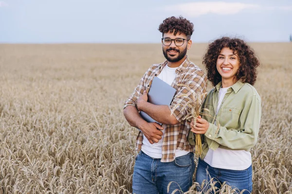 Two agronomists inspecting a wheat crop in a field, holding a laptop and a bundle of wheat ears, showcasing modern agricultural practices