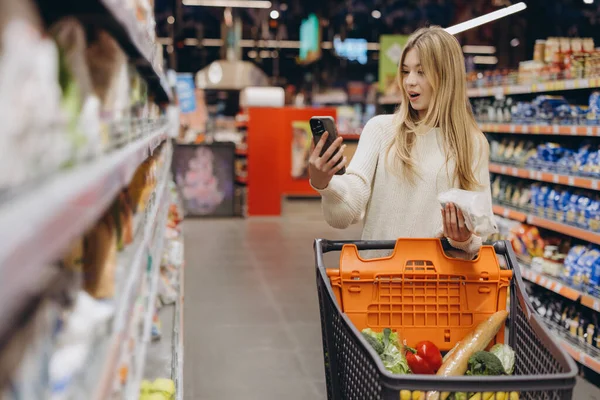 Akıllı telefon kullanan ve marketten alışveriş arabası alan genç bir kadın.