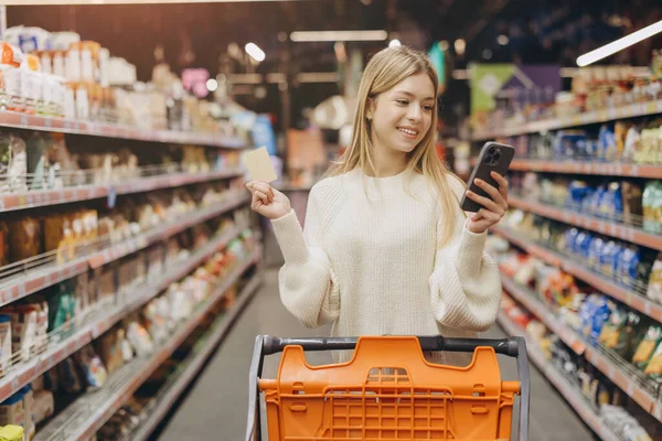 Smiling woman using her phone and credit card while making purchases for groceries in a bright, organized supermarket aisle