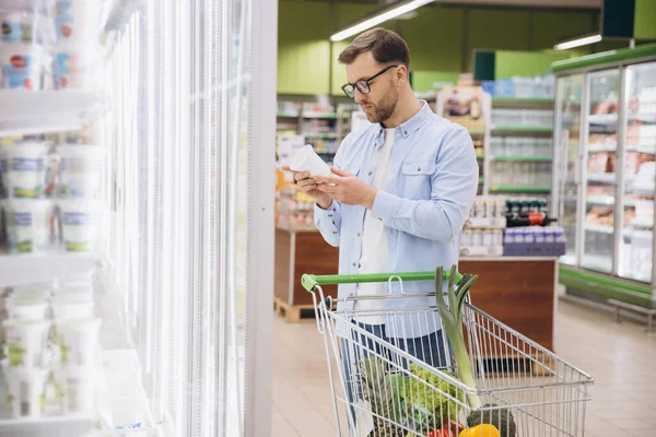 Man pushing shopping cart and reading food label in supermarket refrigerator