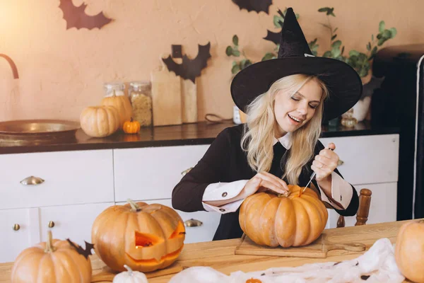 Young woman wearing witch hat carving pumpkin for Halloween party in kitchen decorated with bats and pumpkins