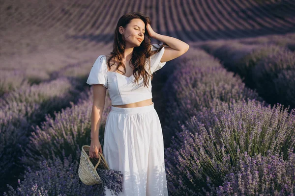Young woman with closed eyes holding a straw bag and a bunch of lavender in a lavender field, enjoying the scent and the summer breeze