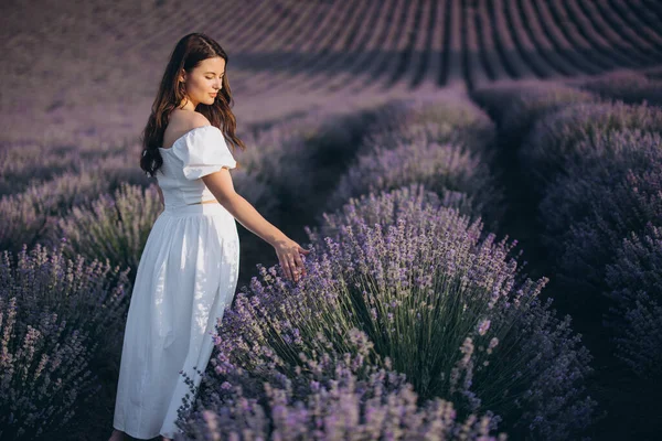 Model wearing a flowing white dress, gently touching vibrant lavender flowers while walking through a serene field under the summer sun