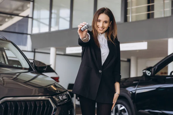Smiling saleswoman holding car keys while standing next to a sleek new car in a modern dealership showroom, exuding confidence and success