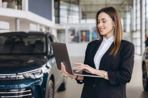 Smiling saleswoman engaging with a laptop while standing beside a new electric car in a modern dealership showroom