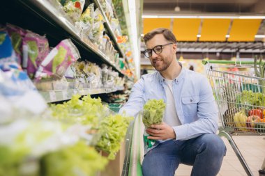 Customer selecting fresh produce in grocery store, holding lettuce and smiling, shopping cart in background
