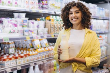 Smiling woman holding milk bottle while shopping in supermarket dairy aisle, choosing healthy products for balanced diet