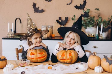 Mother and daughter wearing witch hats, looking at carved pumpkins during Halloween party preparation in decorated kitchen