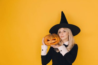 Halloween witch wearing a black hat and dress, joyfully holding a carved pumpkin against a vibrant yellow background